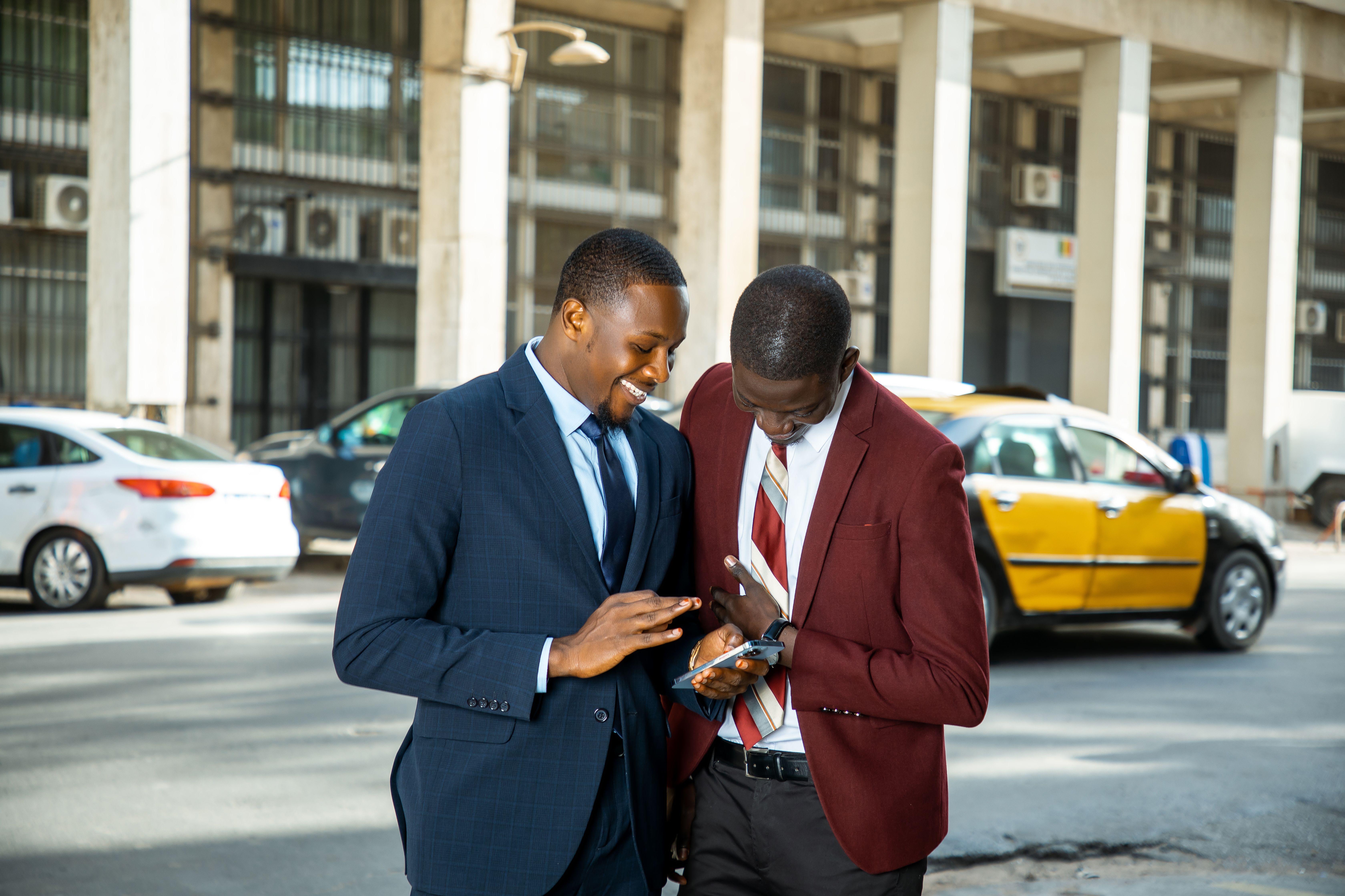 Two businessmen in suits engaging with a mobile phone on a busy street in Dakar. Photo by Libasse El Arbi Ndoye on pexels.