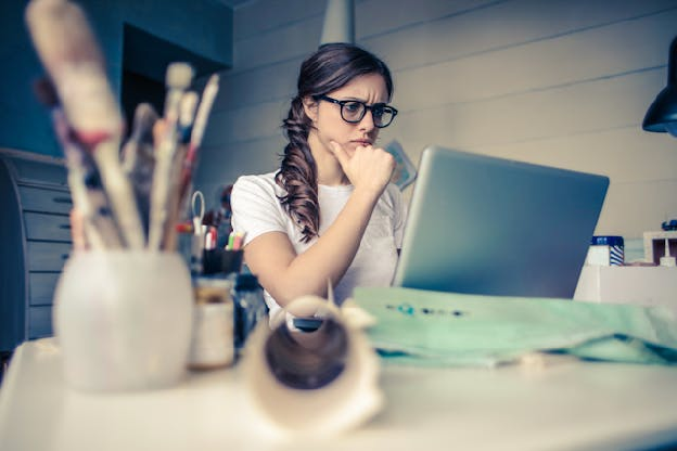 Young woman with glasses deeply focused on a laptop surrounded by art supplies in a home office. Photo by Andrea Piacquadio on pexels.com