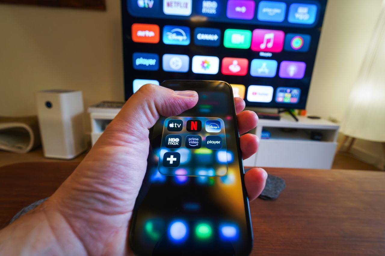 Close-up of a hand holding a phone displaying streaming apps in front of a TV with multiple app icons. Photo by Jakub Zerdzicki on pexels.